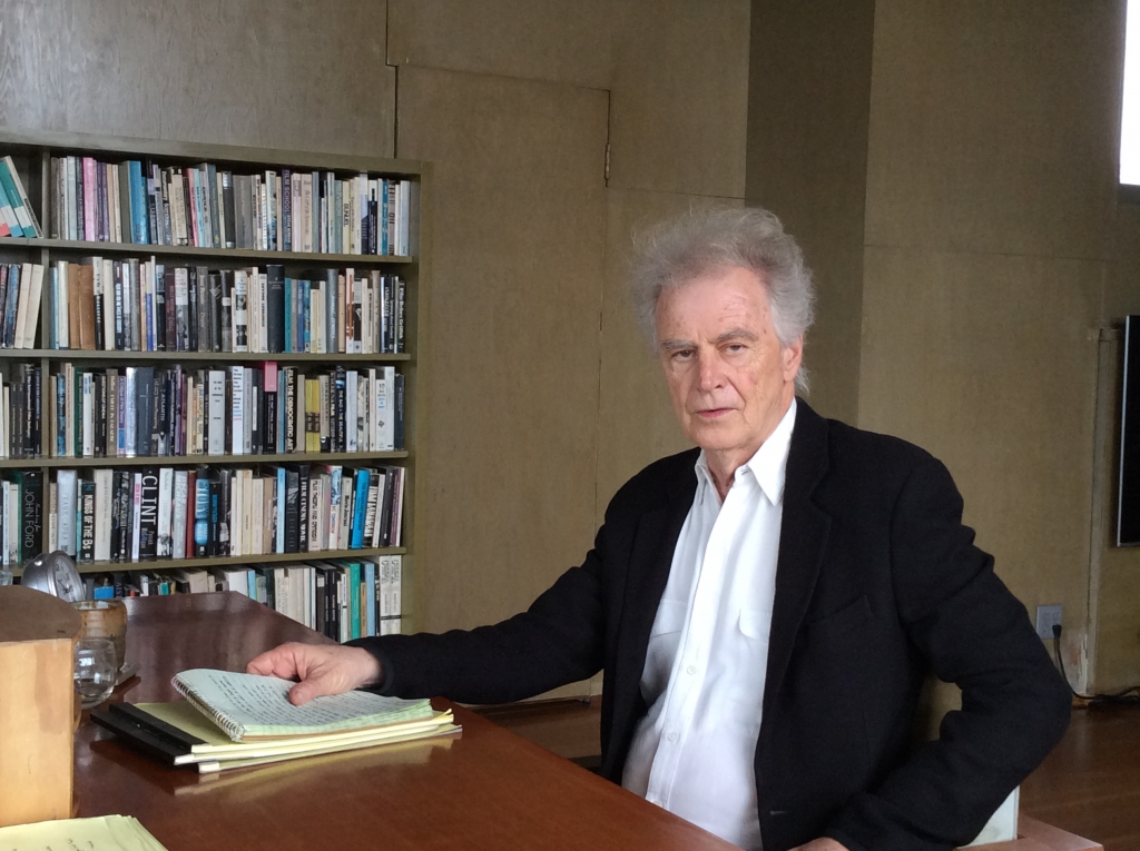 Thom Andersen in a white collared shirt and blue blazer sits at a table with notepads with a bookcase at left.