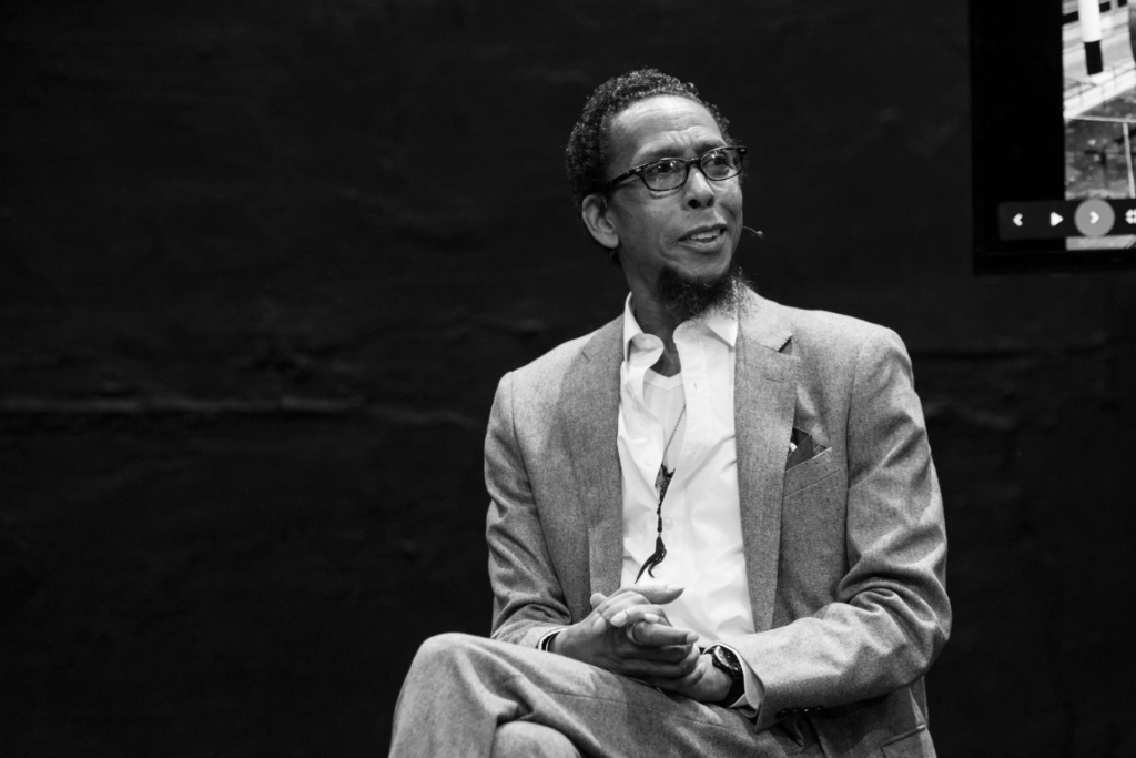 A black and white image of Ron Cephas Jones seated wearing a suit and glasses. 