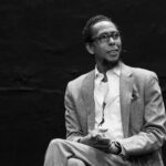 A black and white image of Ron Cephas Jones seated wearing a suit and glasses.