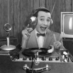 Paul Reubens sits at a desk with headphones and old-school radio and TV equipment as Pee-wee Herman in this black-and-white photo.