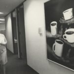 Black-and-white image of person strolling hallway as they look at artwork on wall. The artwork depicts four hands balancing cups of tea or coffee.