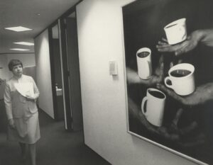 Black-and-white image of person strolling hallway as they look at artwork on wall. The artwork depicts four hands balancing cups of tea or coffee.