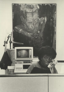 Black-and-white photo of a person answering phone with large painting in the wall behind them.