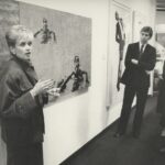 Black-and-white image of Lynne Sowder (left) in front of artwork on the wall, observed by three people on the right.