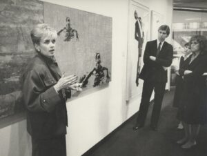 Black-and-white image of Lynne Sowder (left) in front of artwork on the wall, observed by three people on the right.