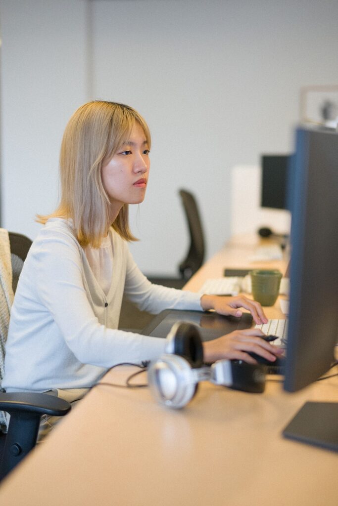 Woman sitting at a computer with a large monitor and headphones on the desk in foreground.