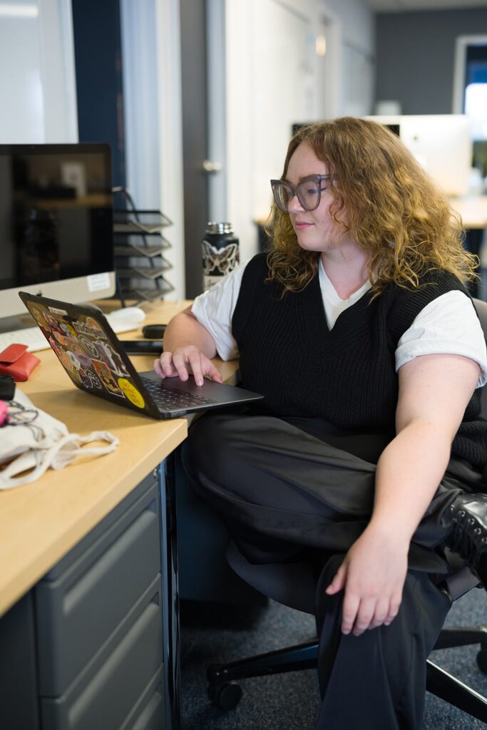 A woman sits at a desk working on a laptop adorned with stickers