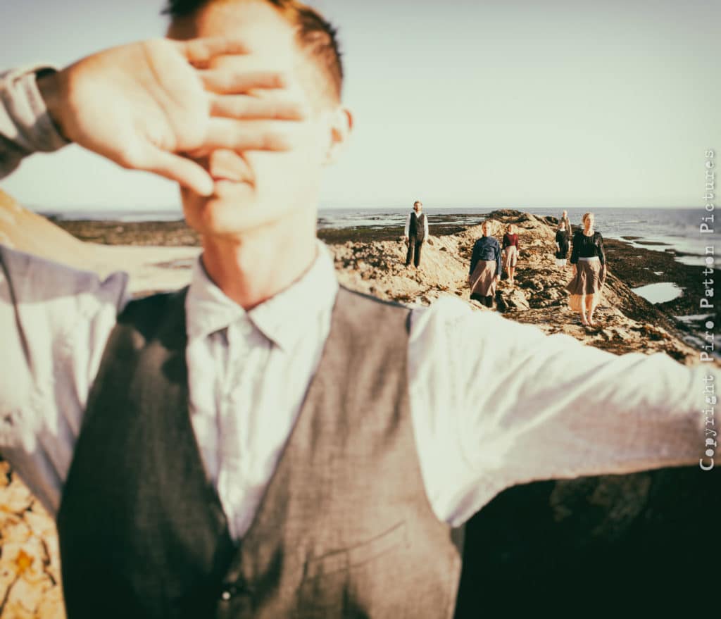 Man covers eyes with hands while others stand in the background against a seascape.