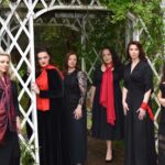 six women dressed mostly in black and red, standing outside under a lattice work