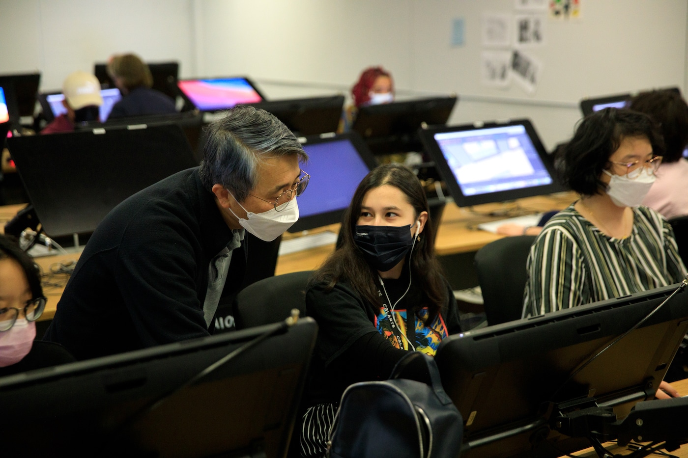 in a computer lab, one faculty member discusses work on a screen with a student