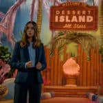 Alison Brie stands in front of a tiki themed TV show set with a sign that reads 'Dessert Island All Stars.'