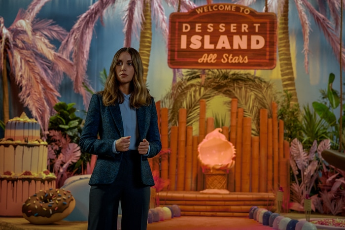 Alison Brie stands in front of a tiki themed TV show set with a sign that reads 'Dessert Island All Stars.'