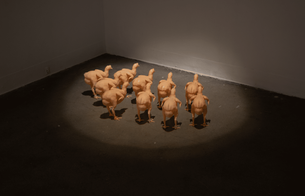 10 plastic chickens, flesh colored, in a spotlight facing a corner wall.
