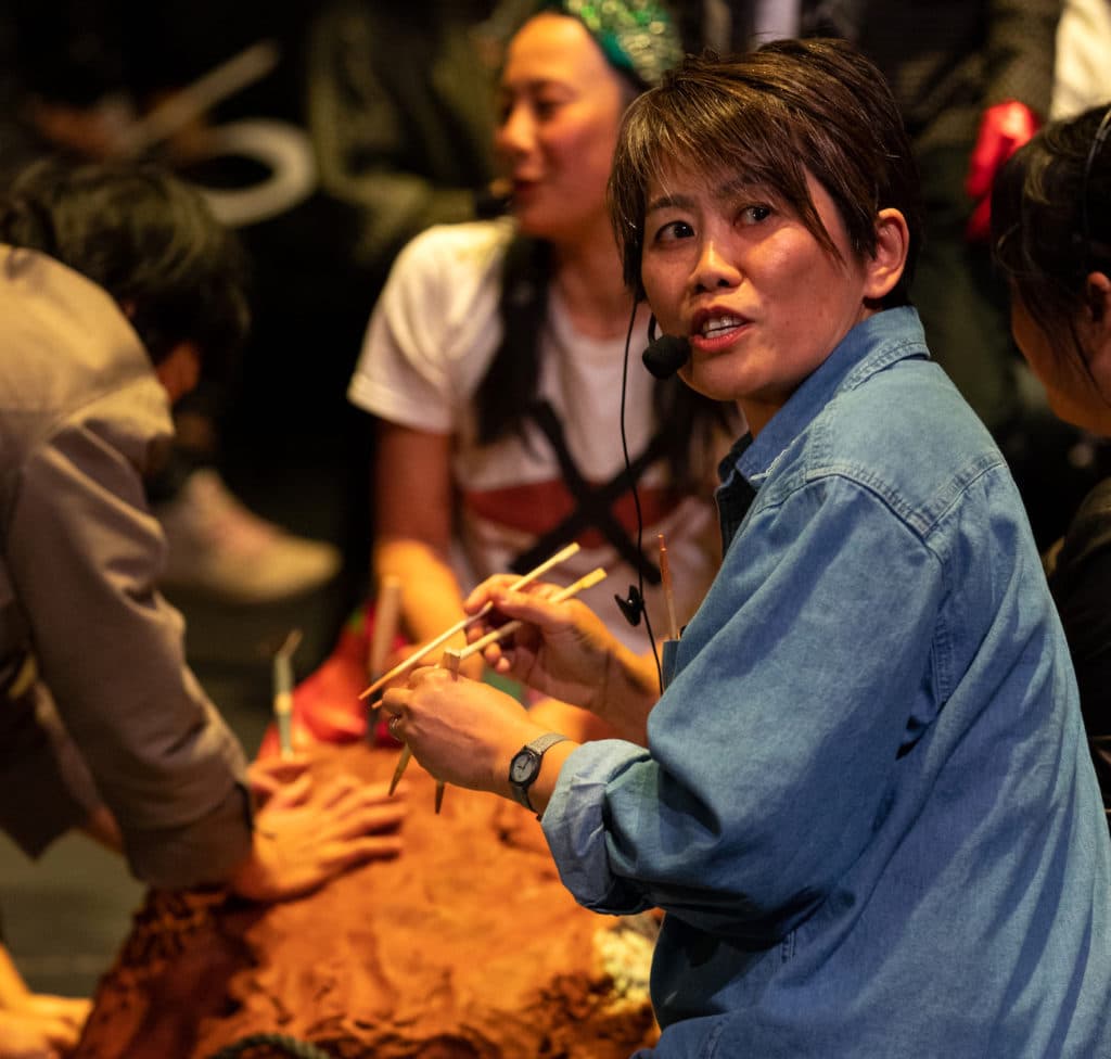 Shirley Tse, kneeling in a blue shirt and chopsticks in her hands, speaks into a headset mic during a performance.