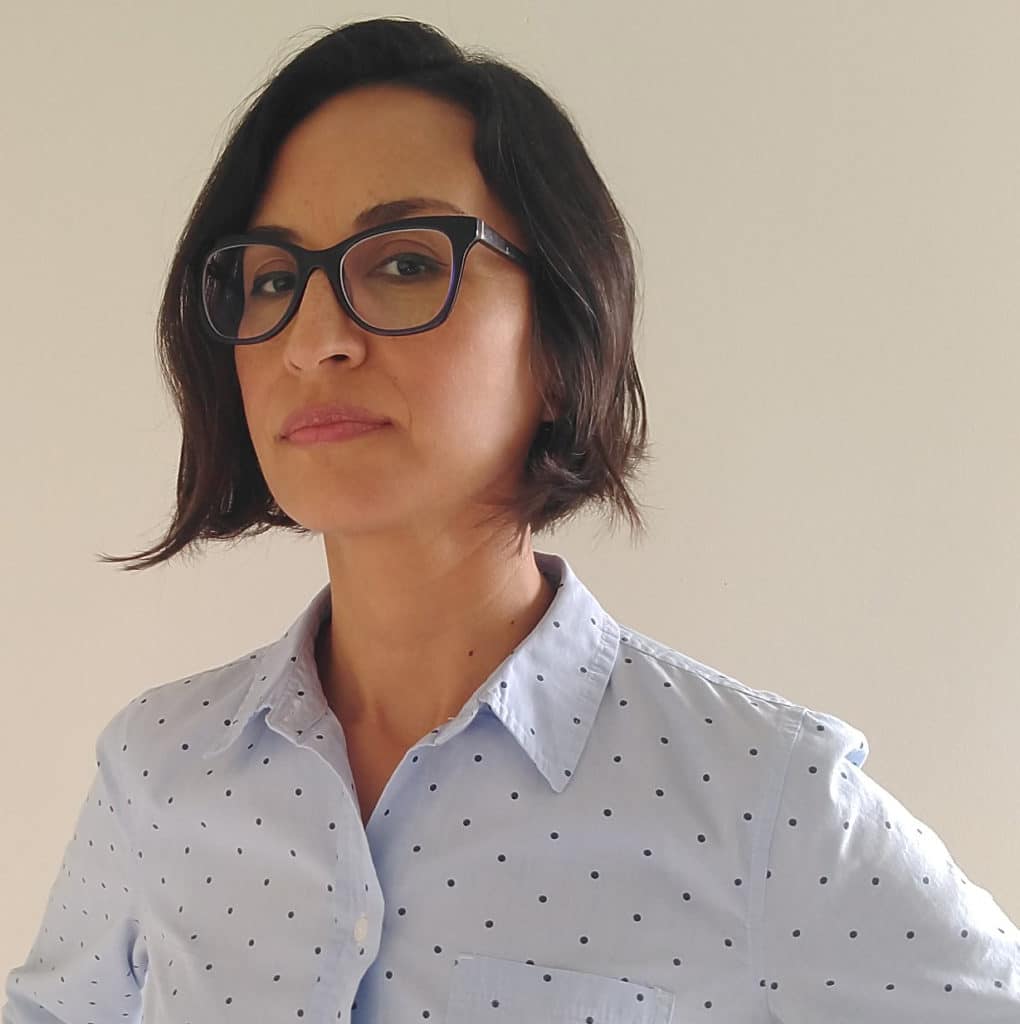 A woman in a collared shirt and glasses glances almost sideways at the camera.
