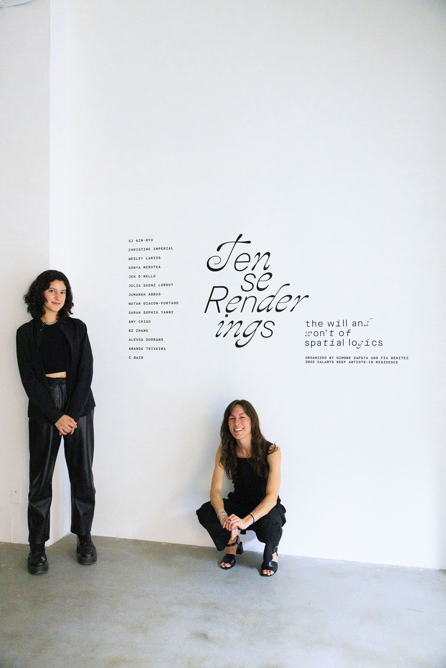 2022 REEF Artists-in-Residence Fía Benitez (standing) and Simone Zapata (crouched) in front of gallery wall featuring 'Tense Renderings' texts and list of artists