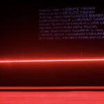 Dancer stands against a dark wall, featuring a projection of names and illuminated with red light