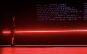 Dancer stands against a dark wall, featuring a projection of names and illuminated with red light