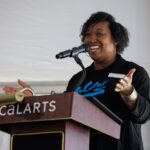 Chevonne Totten-Garner speaks into a mic at a CalArts lectern, wearning a black T-shirt that reads 'CalArts.'