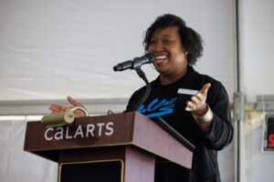 Chevonne Totten-Garner speaks into a mic at a CalArts lectern, wearning a black T-shirt that reads 'CalArts.'