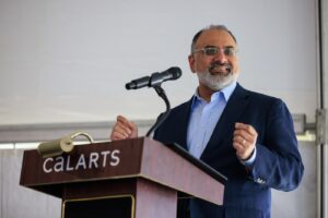 A close up of CalArts President Ravi Rajan standing behind a podium that reads, 'CalArts.' He wears a blue Oxford shirt and a blue sports coat.