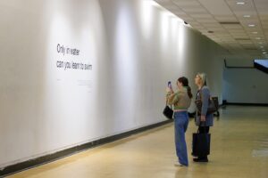 Two women stand in the Main Gallery, one taking a picture of the exhibition title, 'Only in water can you learn to swim.'