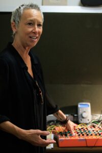 Amy Knoles, in a black shirt, presents a talk with an orange music processor board.