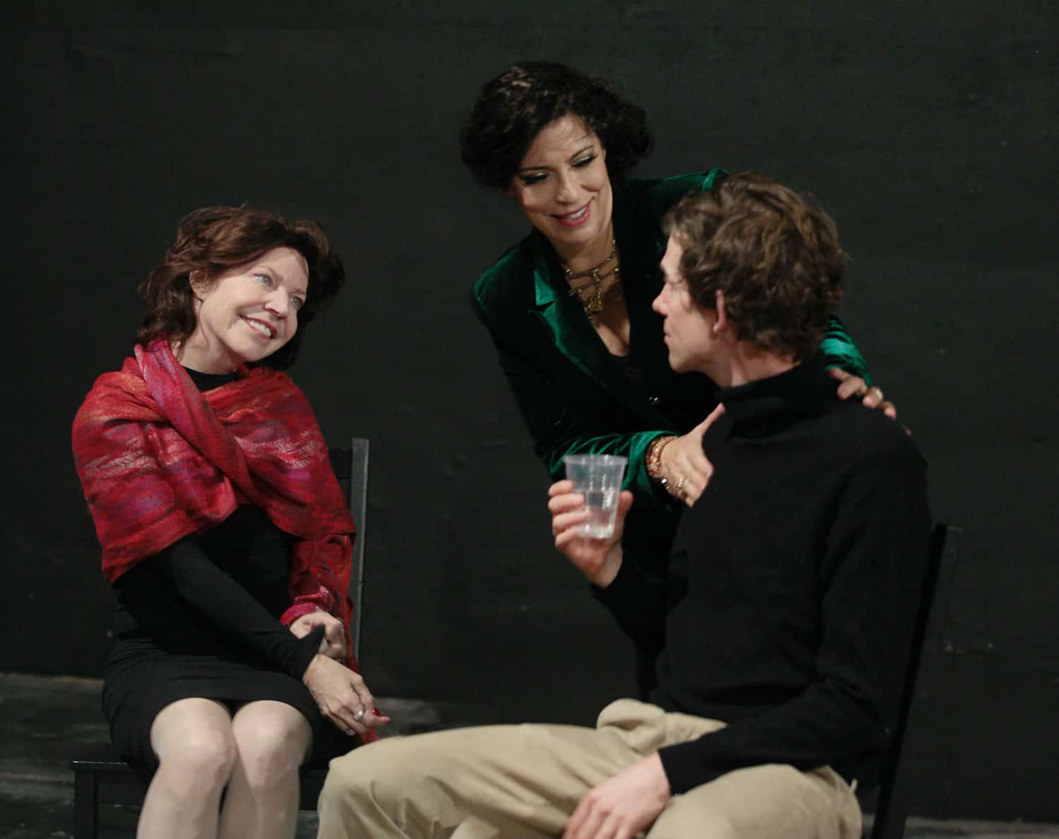Seated woman in red shawl smiles at woman in green velvet blazer, whose hands rest on man in black turtleneck's shoulders