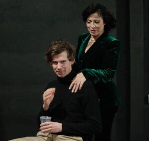 Woman in velvet green blazer rests hands on shoulder of man in black turtleneck.