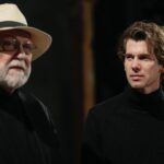 Man in black turtleneck (right) looks contemplatively at bearded man in hat, who gazes into the distance