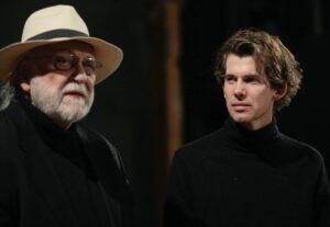 Man in black turtleneck (right) looks contemplatively at bearded man in hat, who gazes into the distance