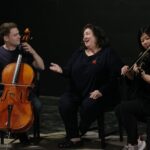 Woman (middle) converses with cellist (left) as violinist (right) plays beside her.