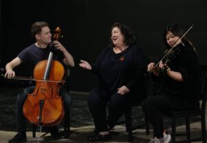 Woman (middle) converses with cellist (left) as violinist (right) plays beside her.