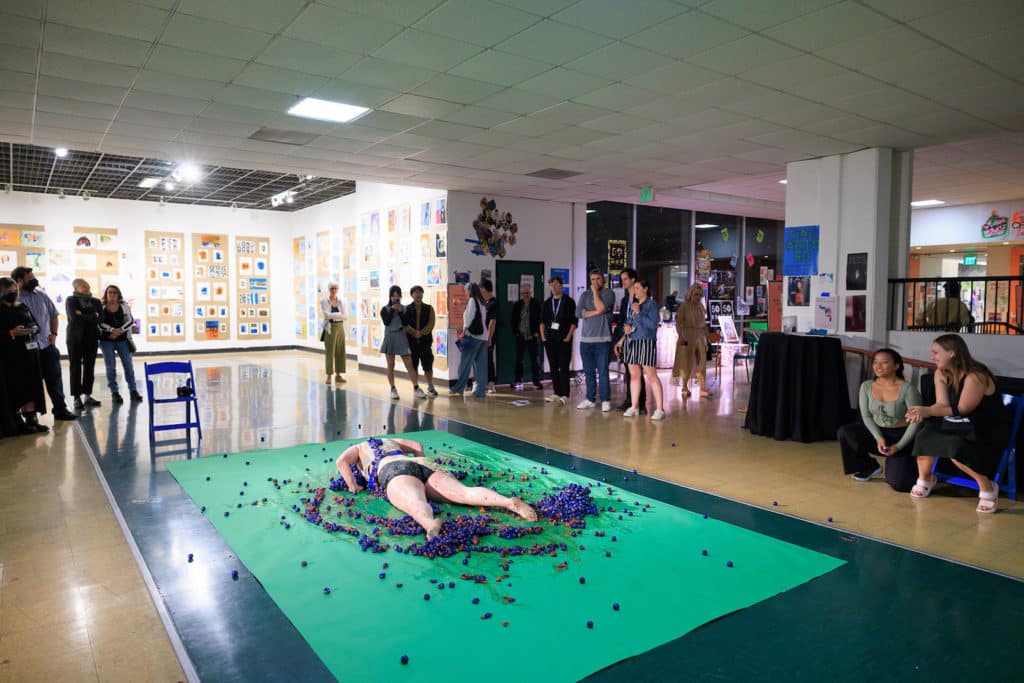 A person lays face down at CalArts on a green tarp on top of tomatoes painted purple while an audience watches.