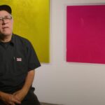 Ruben Ortiz-Torres folds hands in lap, seated in front of gallery wall with bright gold and magenta canvases