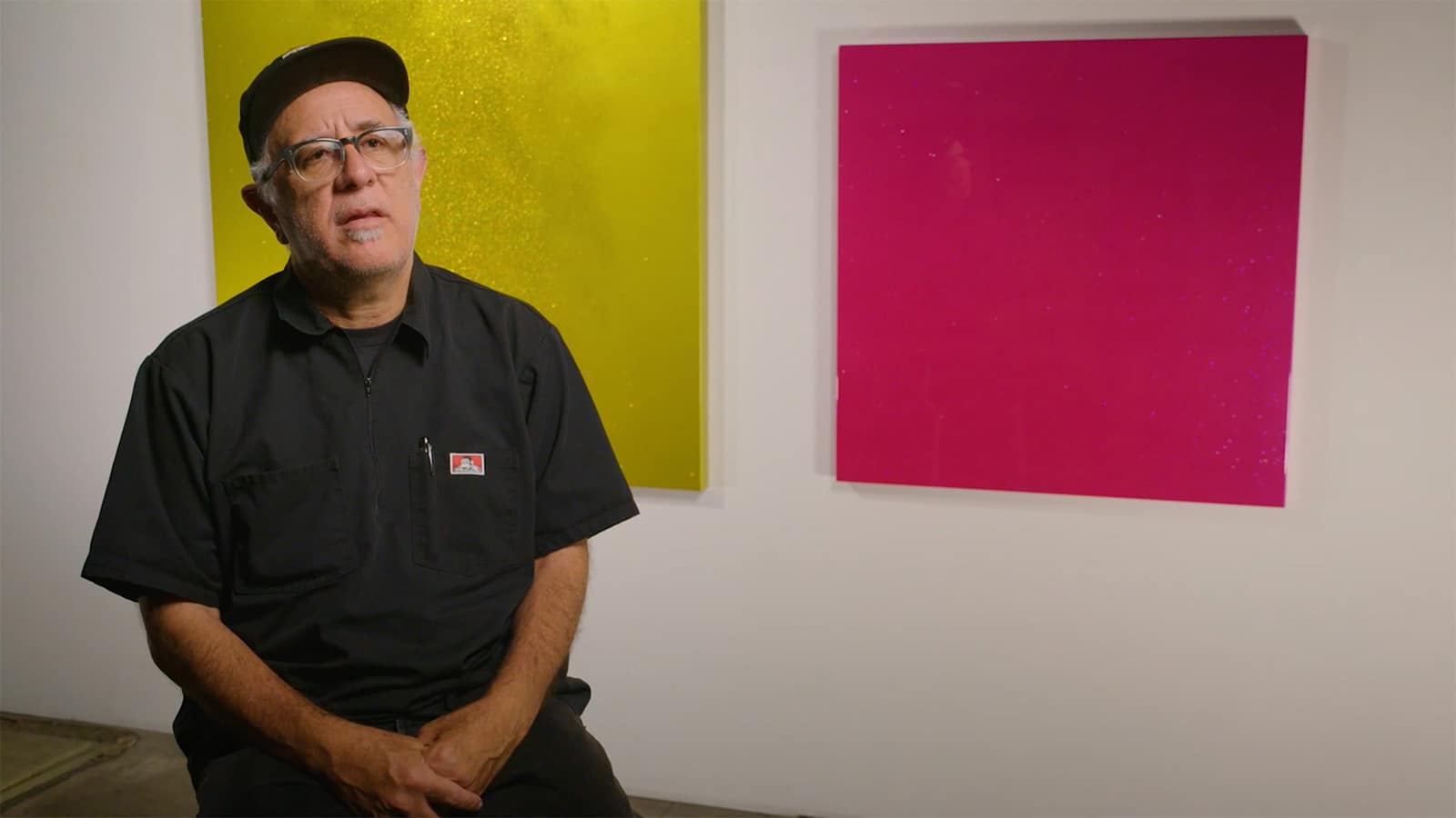Ruben Ortiz-Torres folds hands in lap, seated in front of gallery wall with bright gold and magenta canvases