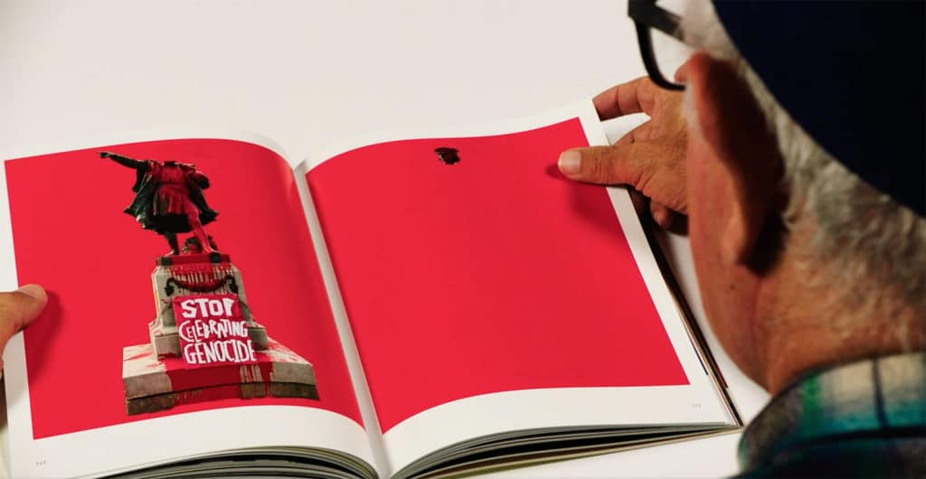 Person holding book spread with bright magenta background and left page with bloodied statue with sign 'Stop Celebrating Genocide'