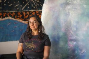 Mercedes Morame standing in front of art in a black t-shirt with wolves that reads, 'Dreamers and Schemers.'