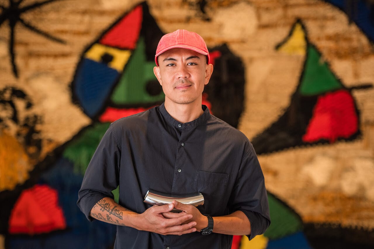 Artist, wearing orange cap and black button-up, holding cylindrical metal object.