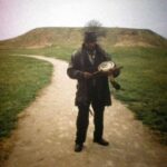Man in hat and overcoat plays drum while standing on a hillside path