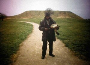 Man in hat and overcoat plays drum while standing on a hillside path