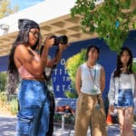 Student standing in front of Blue Wall with long hair and black bandana holds camera as two other students look in camera's direction