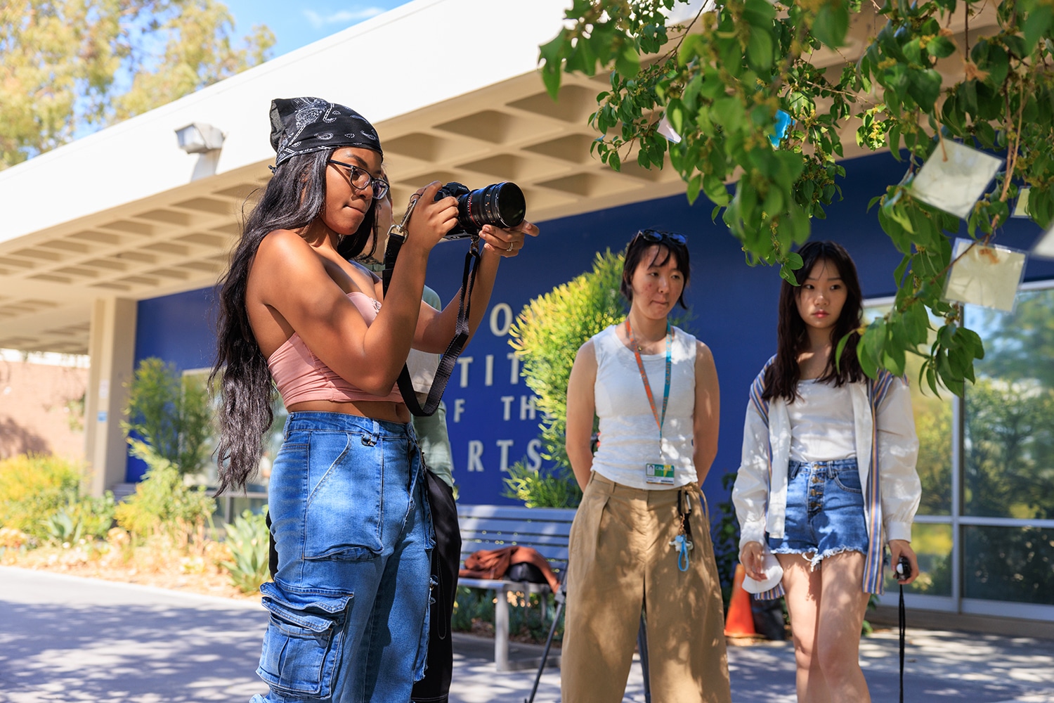 Student standing in front of Blue Wall with long hair and black bandana holds camera as two other students look in camera's direction