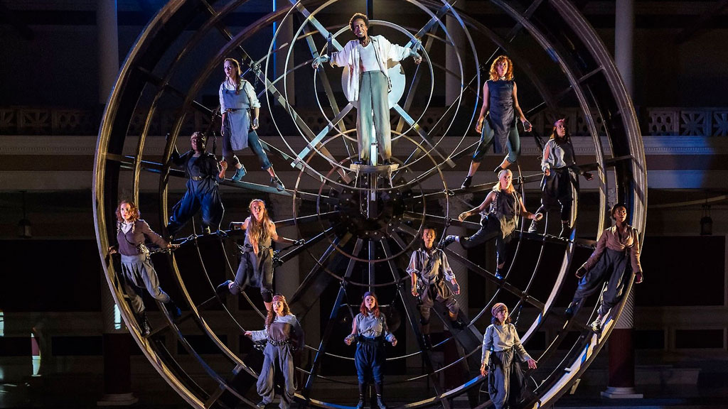A production still from 'Prometheus Bound' with Ron Cephas Jones at center, suspended from a large wheel surrounded by other performers also strapped to the wheel.