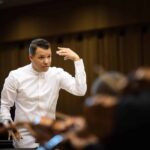 Ryan Bancroft conducts an orchestra. Dressed in a white shirt, his left arm is raised.
