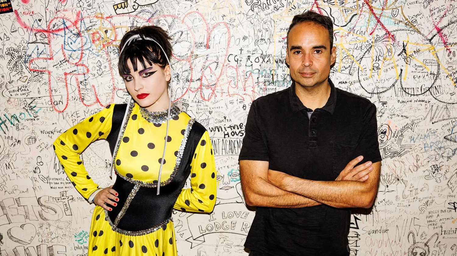 March Adstrum, wearing a yellow polka dot outfit, (L) & John Tejada, wearing a black button-up shirt, (R) stand in front of heavily graffitied wall