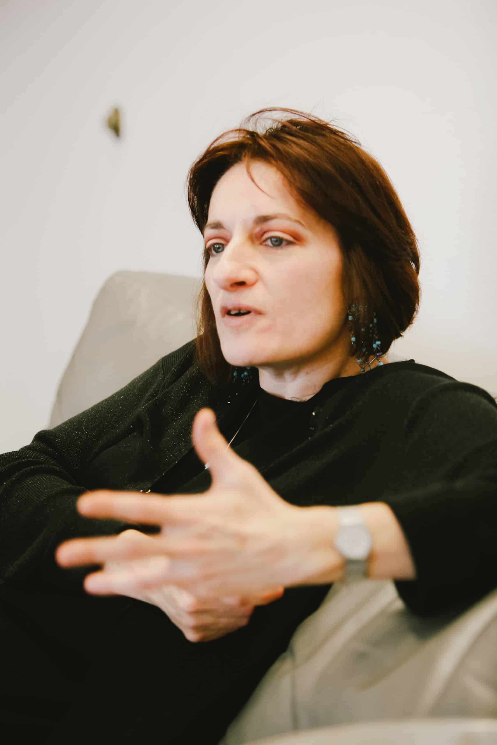 Bérénice Reynaud in a black shirt sits on a beige chair, waving her left hand in the air to accentuate a point.