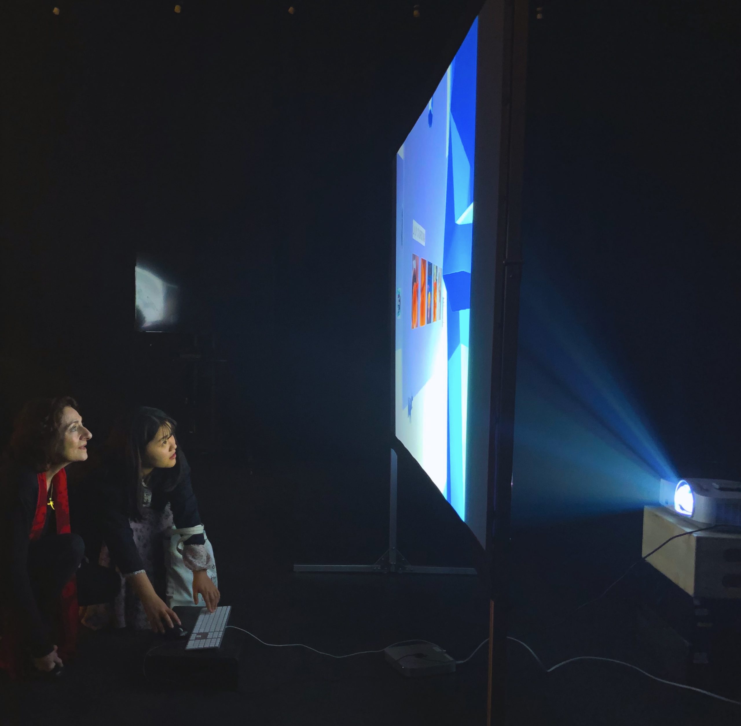 Two women crouch in front of a projected movie screen in a dark room.
