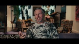 Bill Powers, wearing tie-dye sweater and seated on sofa, gestures with hand while speaking