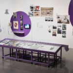 Exhibition view of purple display case filled with images/documents and wall with large purple dots featuring various artworks